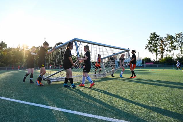 Mannschaft von Fußballspielerinnen hebt ein Tor auf einem Kunstrasenplatz bei Sonnenuntergang an.