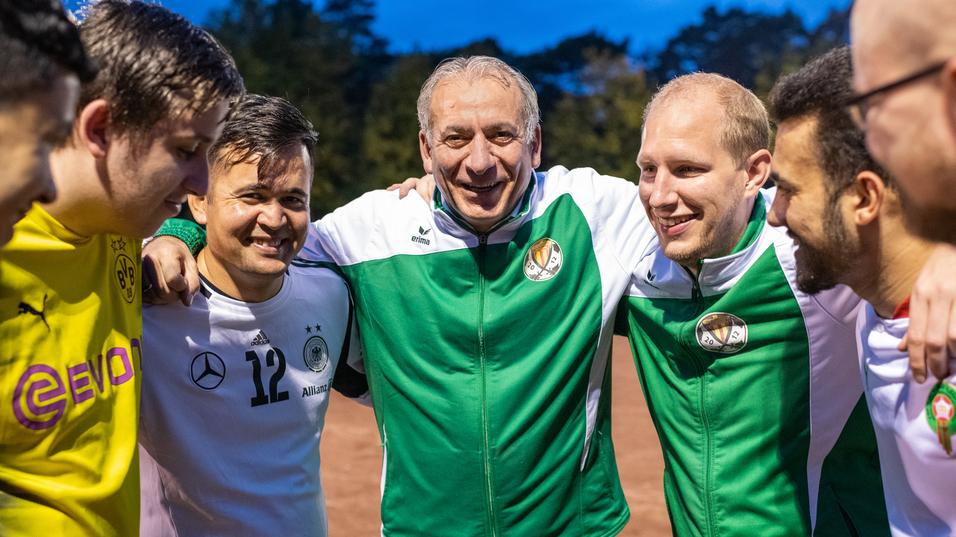 Gruppe von sechs Spielern in Sportkleidung steht lächelnd eng beieinander auf einem Fußballplatz bei Dämmerung.