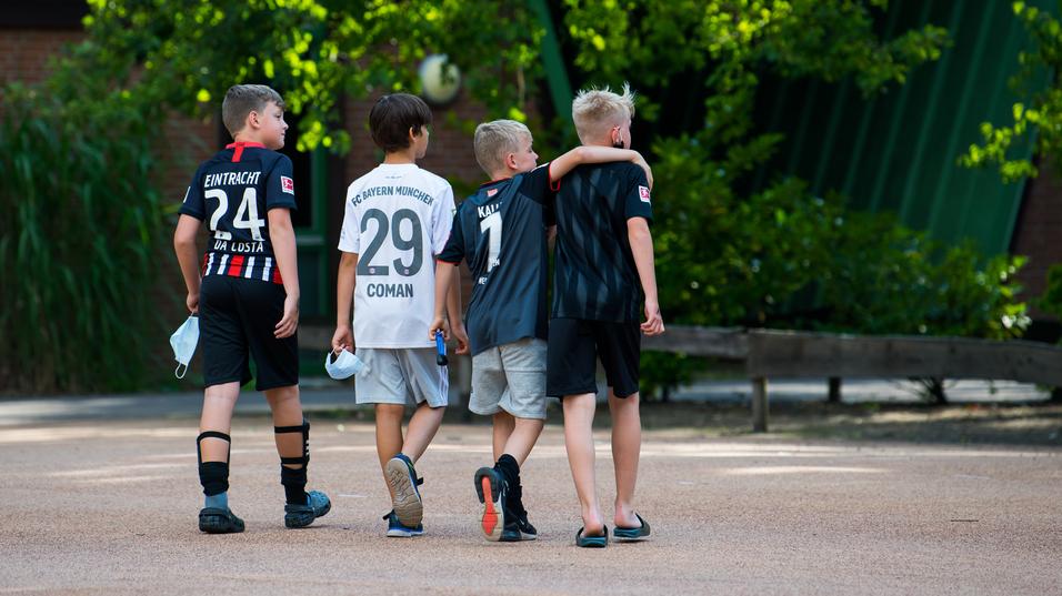 Vier Kinder in Fußballtrikots gehen Arm in Arm über einen Platz, umgeben von Bäumen und einer ruhigen Atmosphäre.