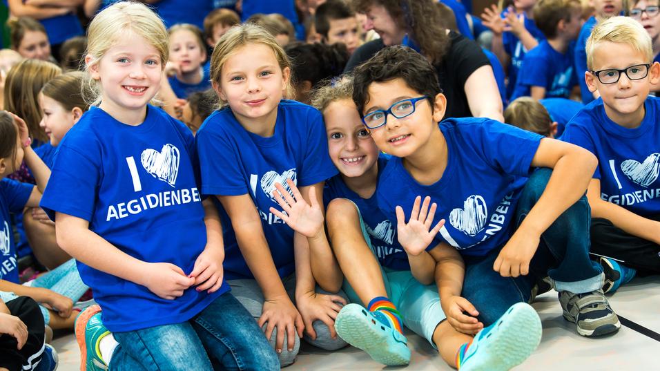 Vier Kinder in blauen T-Shirts mit dem Aufdruck "I ❤️ AEGIDIENBE" posieren lächelnd auf einem Boden.