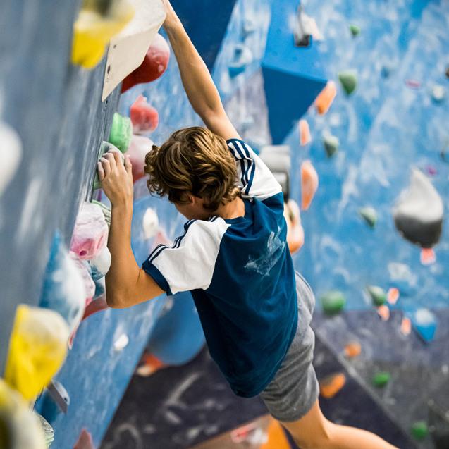 Jugendlicher klettert an einer blauen Boulderwand mit bunten Griffen, schwebt in einer dynamischen Position.