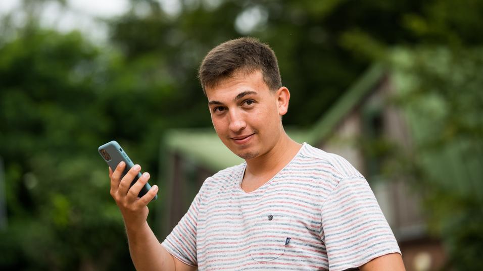 Junger Mann mit kurzen Haaren hält ein Smartphone in der Hand und schaut lächelnd in die Kamera, Hintergrund unscharf.