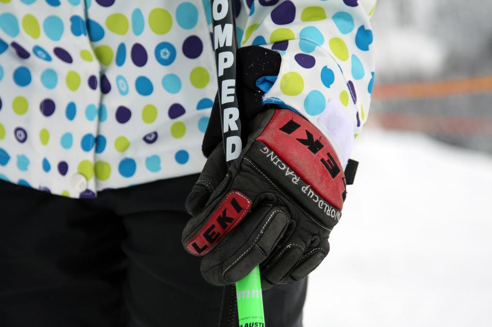 Nahaufnahme einer Hand in einem schwarzen Skihandschuh, die einen Skistöck mit bunten Punkten in einer weißen Jacke hält.