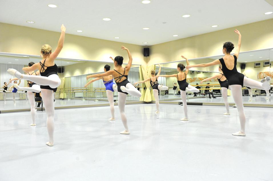 Gruppe von Balletttänzerinnen in Schwarz und Blau übt in einem Spiegelraum mit Holzballettstangen.