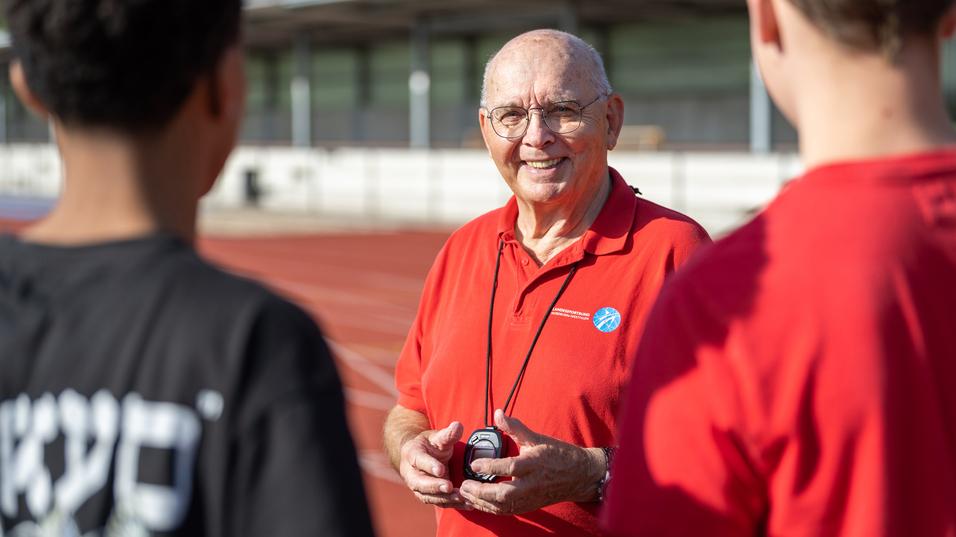 Ein älterer Mann in rotem Shirt mit Stoppuhr spricht an einer Laufbahn mit zwei Jugendlichen.