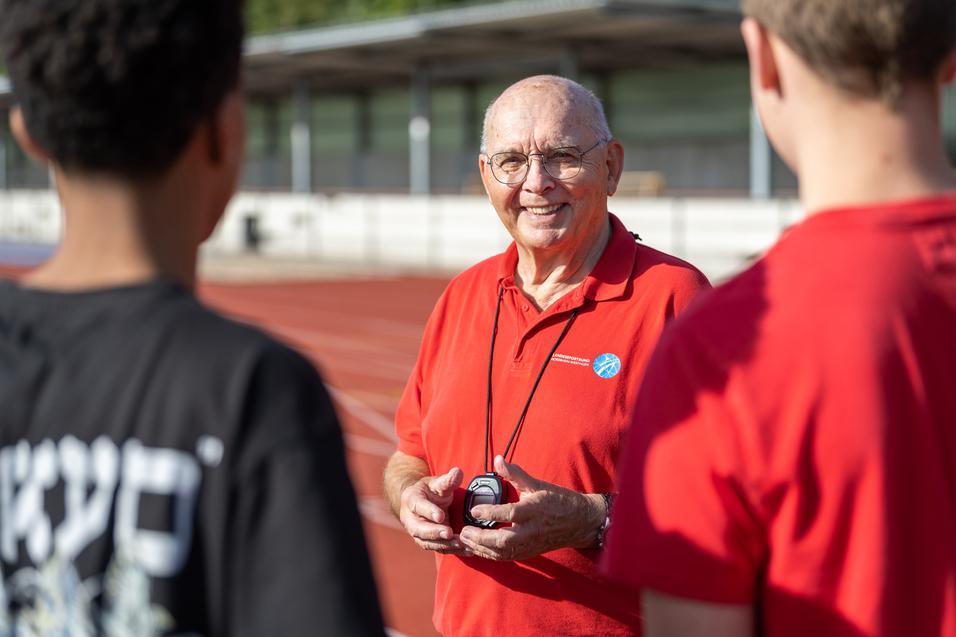 Ein älterer Mann in rotem Shirt mit Stoppuhr spricht an einer Laufbahn mit zwei Jugendlichen.