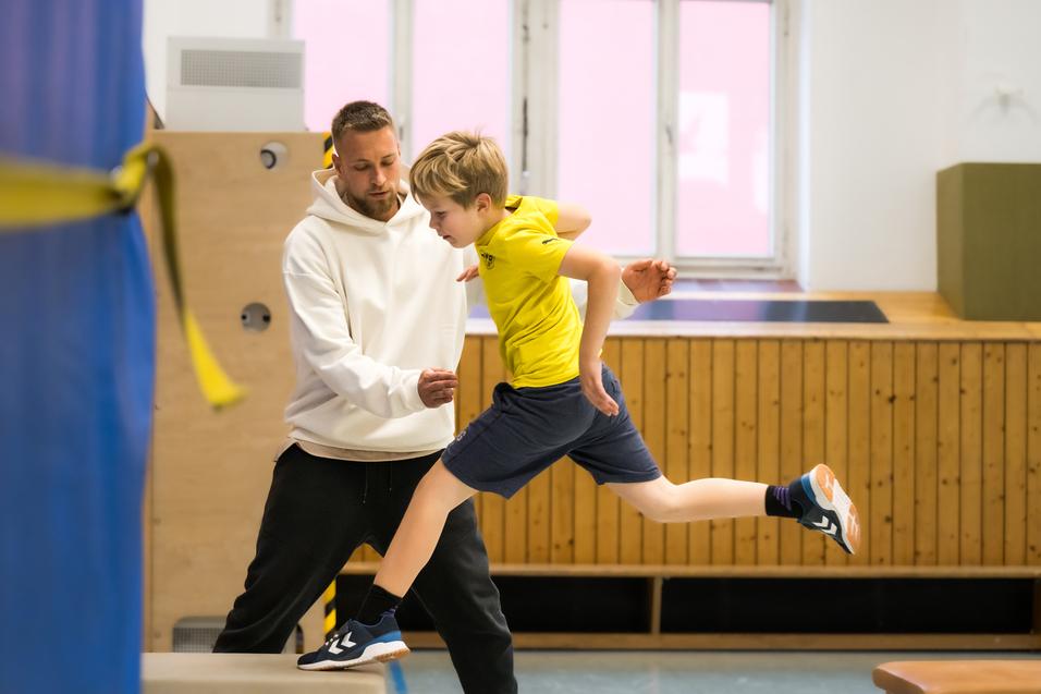 Trainer unterstützt einen Jungen beim Sprung über ein Trainingsgerät in einer Turnhalle.